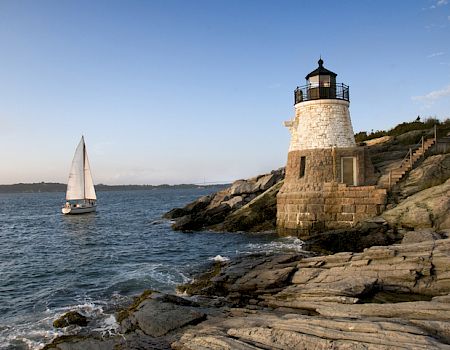 A lighthouse stands on a rocky shore, overlooking a sailboat on the ocean, under a clear sky with gentle waves hitting the rocks.