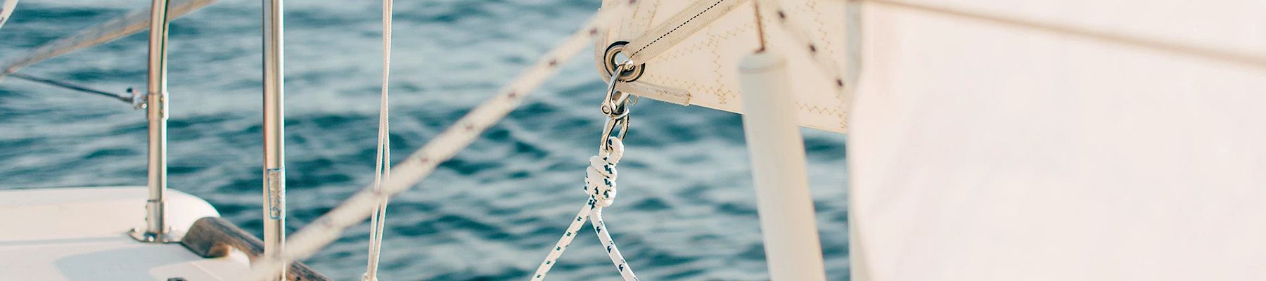 The image shows part of a sailboat with white sails, ropes, and stainless steel railing, set against a backdrop of blue, calm water.