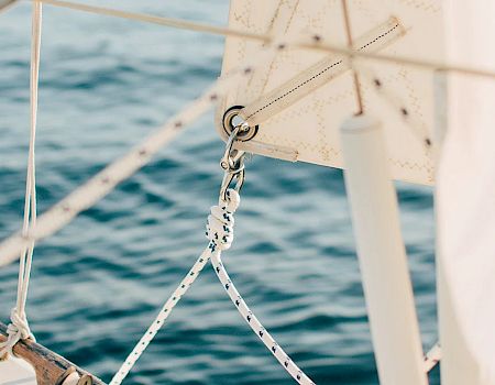 The image shows part of a sailboat with white sails, ropes, and stainless steel railing, set against a backdrop of blue, calm water.