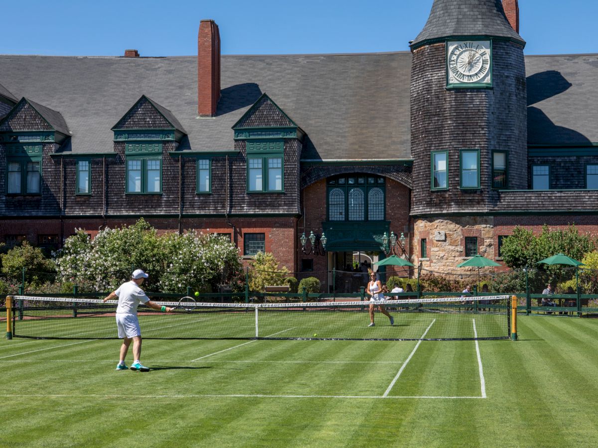 Two people play tennis on a manicured lawn in front of a large, old stone building with a clock tower; a net spans the yard.