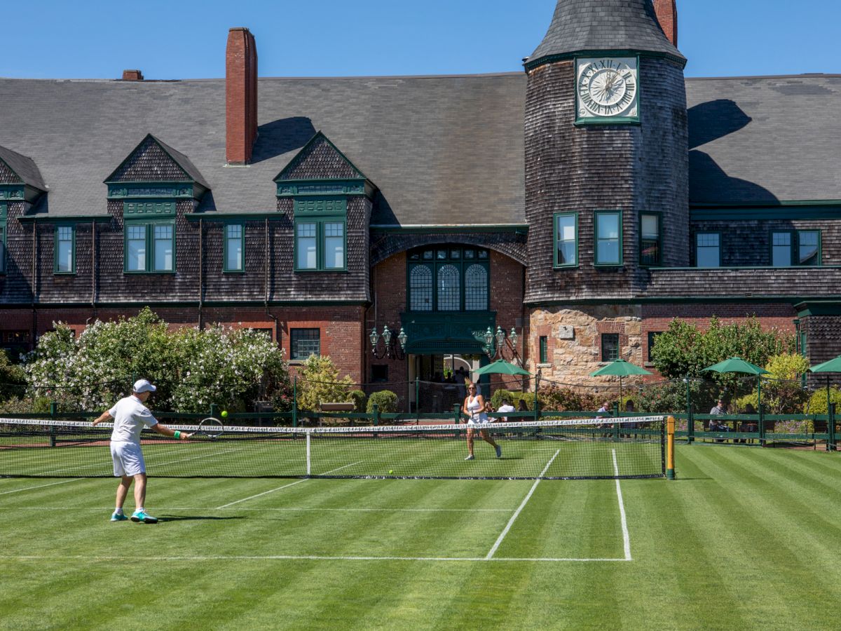 Two people play tennis on a sunny lawn in front of a large stone building with a clock tower, neatly trimmed grass, and a hedge border.
