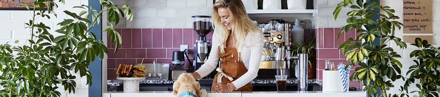 A woman stands behind a blue counter inside a cozy, plant-filled cafe. A dog stretches up to her on its hind legs. The cafe decor is inviting.