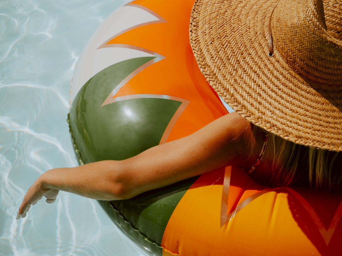 A person floats on a colorful inflatable ring in a pool, wearing a wide straw hat, palm-tree vibes, sunlit water.
