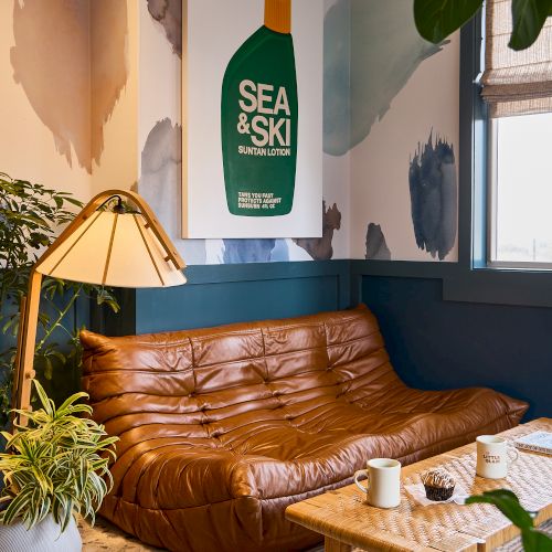 A cozy living room with a brown leather sofa, a wooden coffee table, a plant, and a framed Sea & Ski poster on a paint-splashed wall.