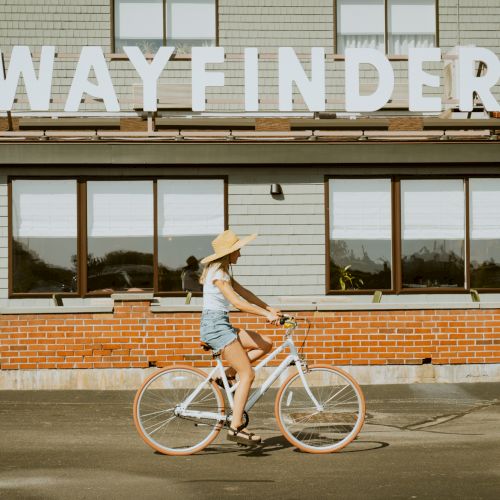 A cyclist rides past a wooden storefront with a sign &ldquo;WAYFINDER&rdquo; on a sunny day.