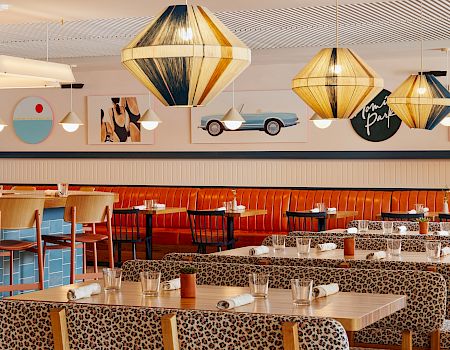 This image shows a stylish restaurant with modern hanging lamps, leopard print seating, and a bar area. Artwork decorates the walls.