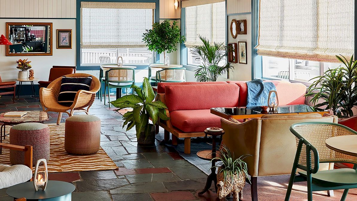 A cozy, modern living space with colorful furniture, various plants, and wall decor. Large windows let in natural light, enhancing the vibrant decor.