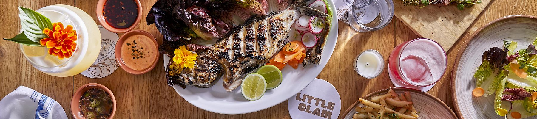 A table spread of grilled fish and meats, fries and salad, plus sauces and drinks, all served family-style.