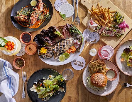 A table spread of grilled fish and meats, fries and salad, plus sauces and drinks, all served family-style.