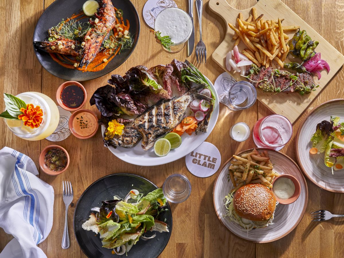 An overhead spread of grilled meats, fish, fries, salad, lime wedges, dipping sauces, and drinks on a wooden table.