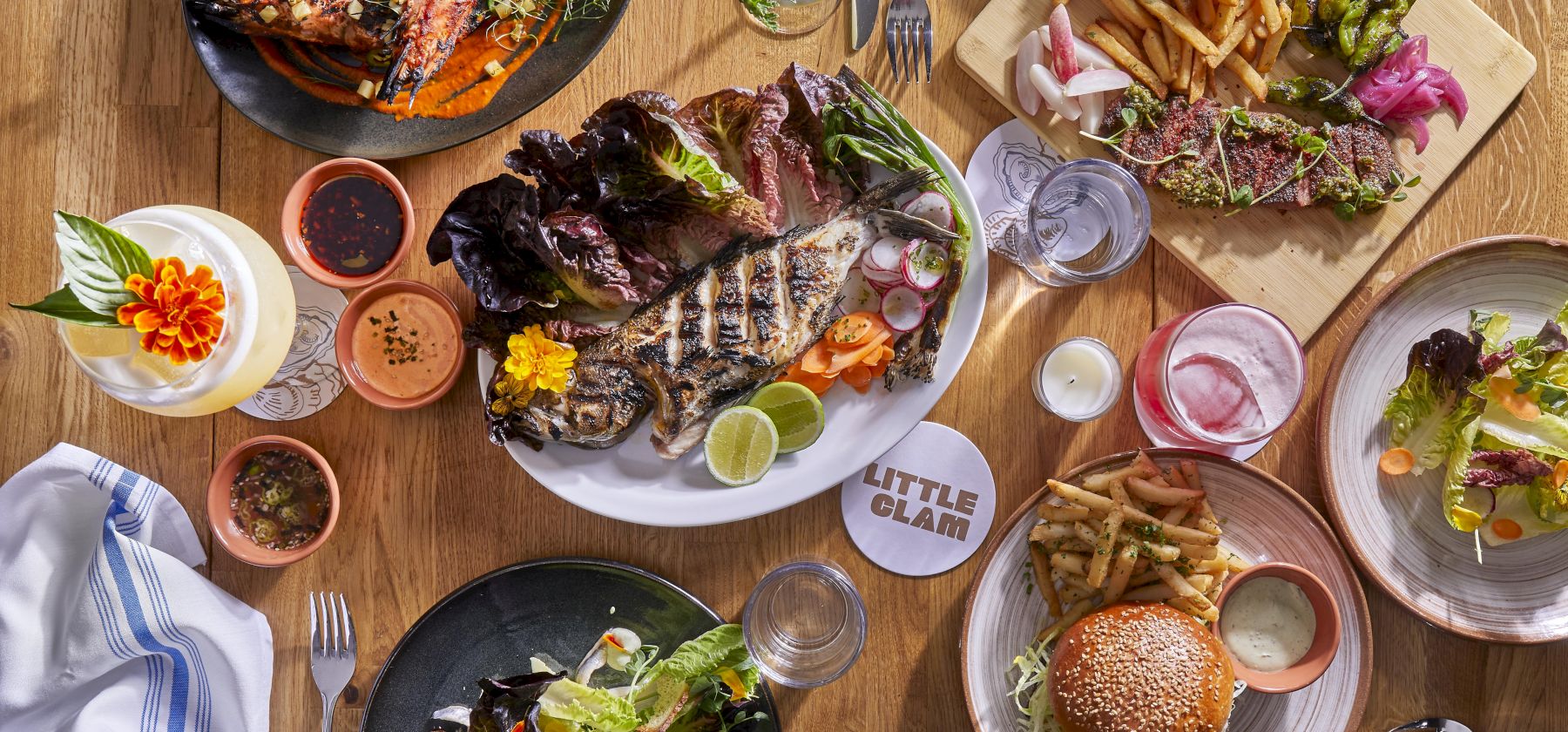 An overhead spread of grilled meats, fish, fries, salad, lime wedges, dipping sauces, and drinks on a wooden table.
