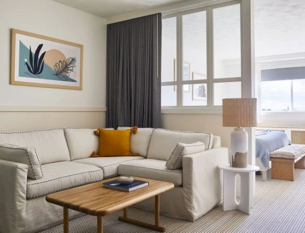 A cozy living area with a neutral-toned sectional sofa, a wooden coffee table, a table lamp, framed art, and a glimpse of a bedroom through a partition.