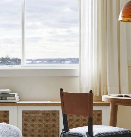 A cozy room with a window, table, chair, and books. A glass of beverage and lamp are on the table, and there's a scenic view outside the window.