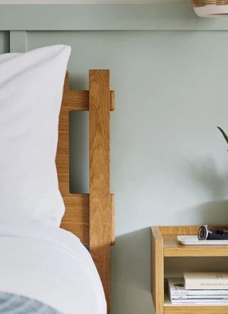 The image shows a neatly made bed with white linens, a wooden headboard, and a nightstand holding a potted plant, books, and a pair of glasses.