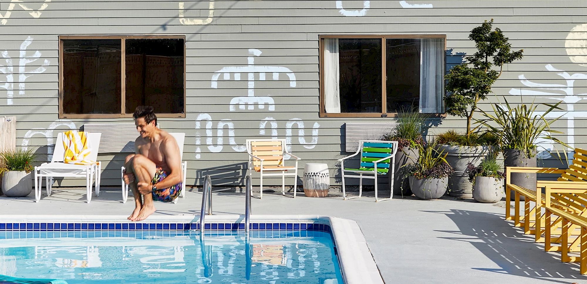 A man in swim trunks crouches by a pool surrounded by colorful chairs and plants, with a building featuring window decor in the background.