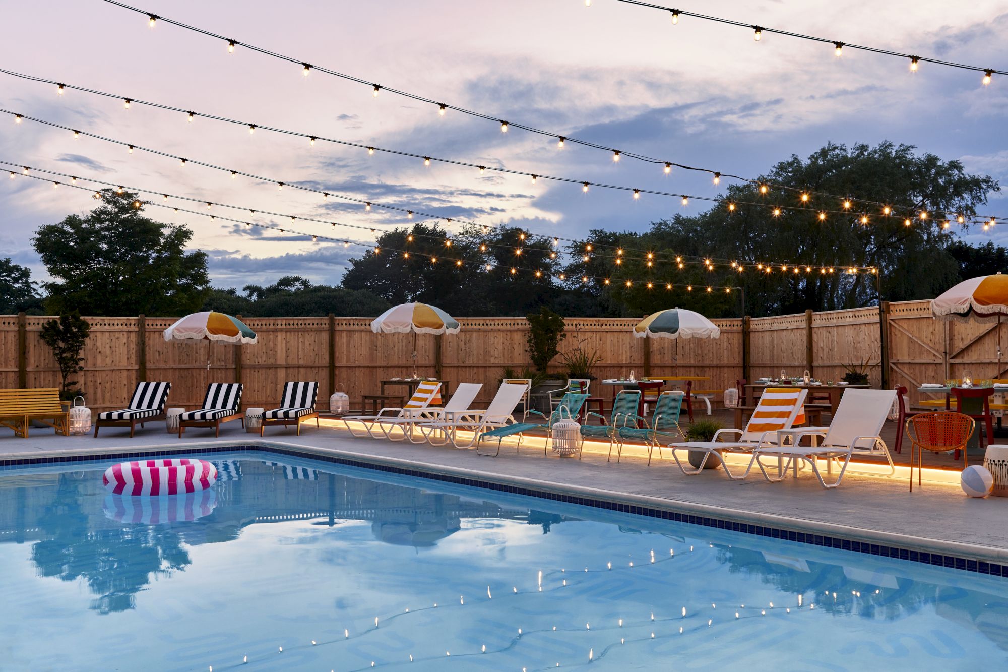 A pool with lounge chairs, string lights, and umbrellas along the deck; calm water reflecting lights under a blue sky.