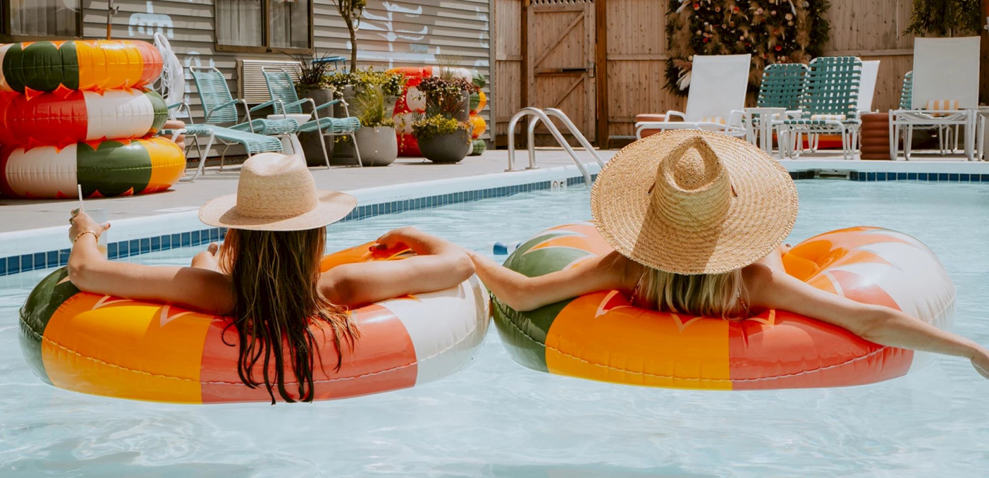 Two people are floating on inflatable rings in a pool, wearing wide-brimmed hats and relaxing. The poolside has chairs and stacked inflatables.