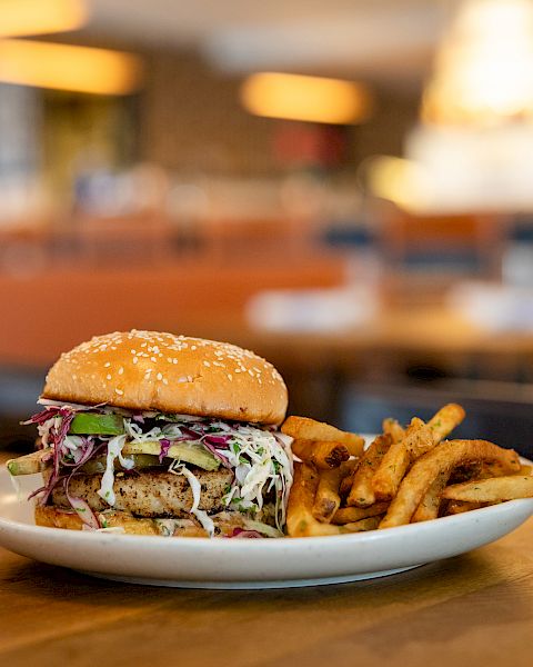 A juicy burger with coleslaw and pickles sits on a plate with crispy fries in a warmly lit restaurant dining room.