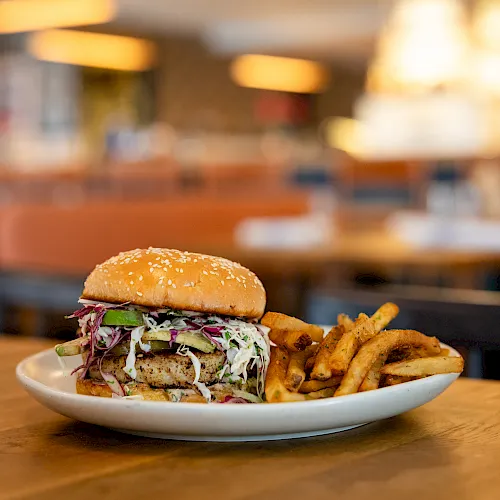 A juicy burger with coleslaw and pickles sits on a plate with crispy fries in a warmly lit restaurant dining room.