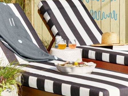 Outdoor patio lounge with striped chaise cushions, drinks on a small table, and a wooden wall painted with blue patterns and a sun.