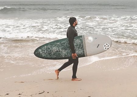 A person carrying a surfboard walks on the beach towards the ocean waves, wearing a wetsuit.