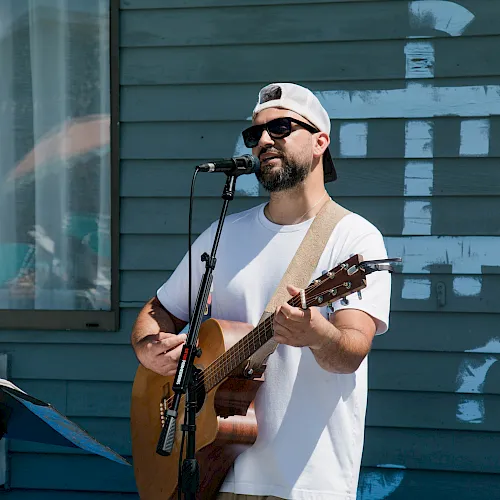A man in a cap and sunglasses performs with an acoustic guitar at a microphone outdoors against a blue building wall.