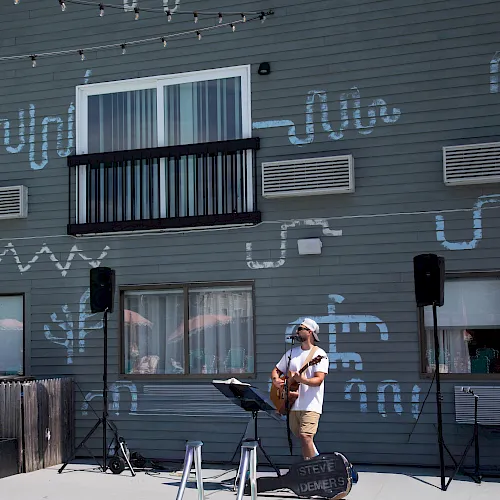 An outdoor music setup with a guitarist at a microphone and speakers, in front of a blue clapboard building covered in graffiti.