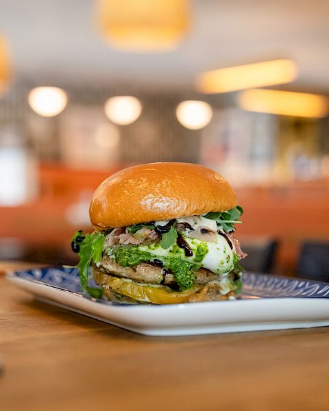 A juicy burger with lettuce and sauce sits on a plate in a warmly lit restaurant, with fries in the background.
