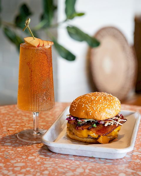 A burger sits on a tray beside a tall orange cocktail garnished with apple slices and a gold pinwheel on a table.