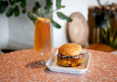 A burger sits on a tray beside a tall orange cocktail garnished with apple slices and a gold pinwheel on a table.