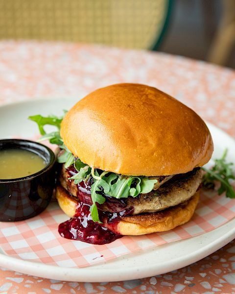 A cheeseburger on a plate with leafy greens, beet/berry sauce, and a small cup of green dipping sauce.