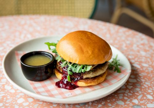 A cheeseburger on a plate with leafy greens, beet/berry sauce, and a small cup of green dipping sauce.
