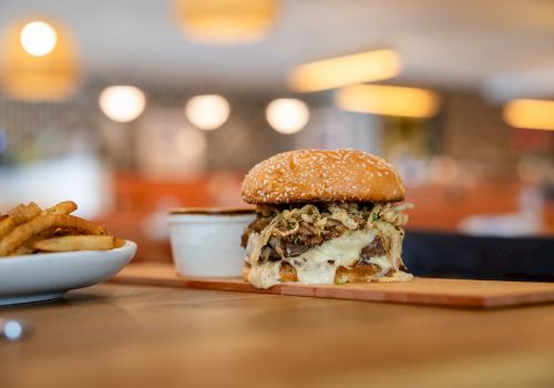 A juicy burger with melted cheese and toppings on a wooden board, with French fries in a bowl in a warm-lit restaurant.