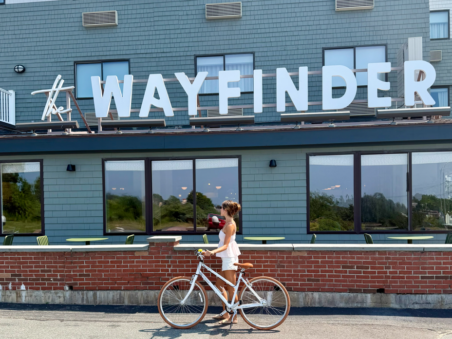 A woman rides a white bike in front of a building with a large &ldquo;ALWAYFNDER&rdquo; sign and windows and air conditioners above, always.