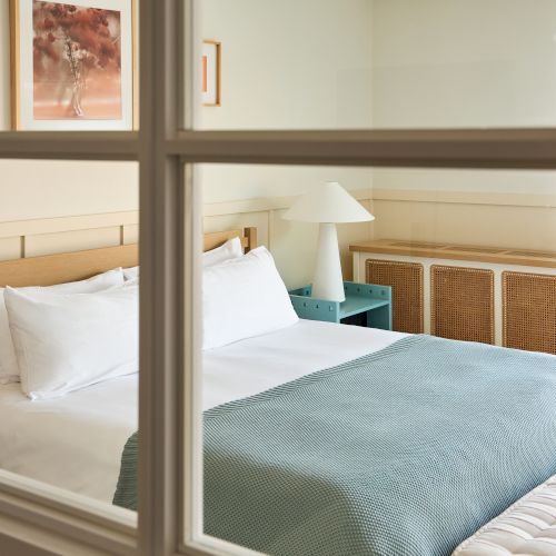 A bright hotel bedroom seen through a window frame, with made beds, a bedside lamp, and framed wall art.