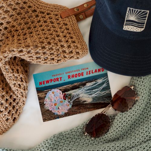 Crocheted scarves/blankets, a navy cap with a sunset logo, a &ldquo;Newport, Rhode Island&rdquo; postcard, and sunglasses laid out.