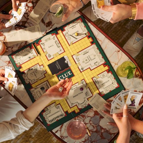 People play Clue on a tabletop, holding suspect cards and a die over the board.