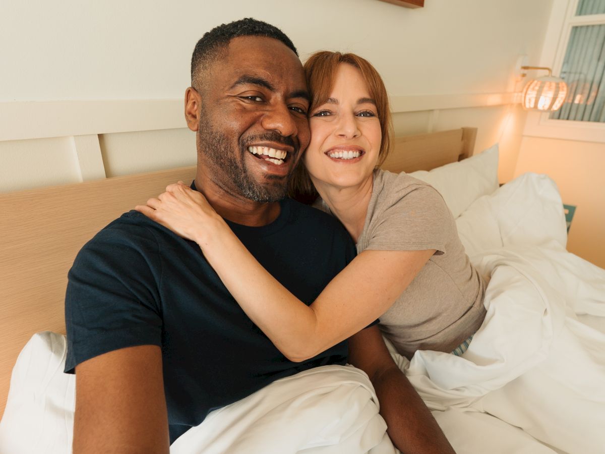 Smiling couple cuddled in bed in a cozy room, with warm lamp light and wall art above them.