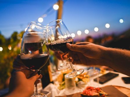 Friends clink wine glasses at a sunny outdoor dinner, toasting under glowing lights and a clear blue sky.