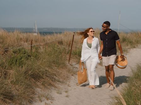 A couple strolling along a sandy beach, carrying woven baskets, smiling and holding hands under a clear sky.