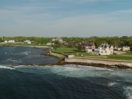 A coastal estate with manicured lawns and white buildings beside a rocky shoreline, waves crashing under a cloudy sky.