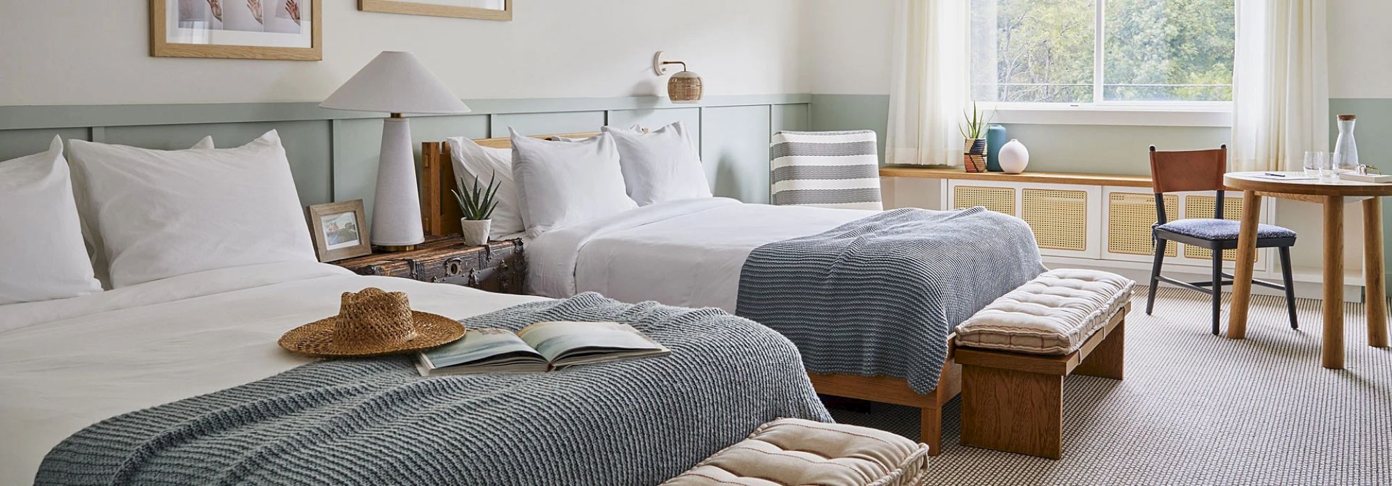 A cozy bedroom with two double beds, soft blue blankets, and white pillows, decorated with books, a hat, and a table by the window with natural light.