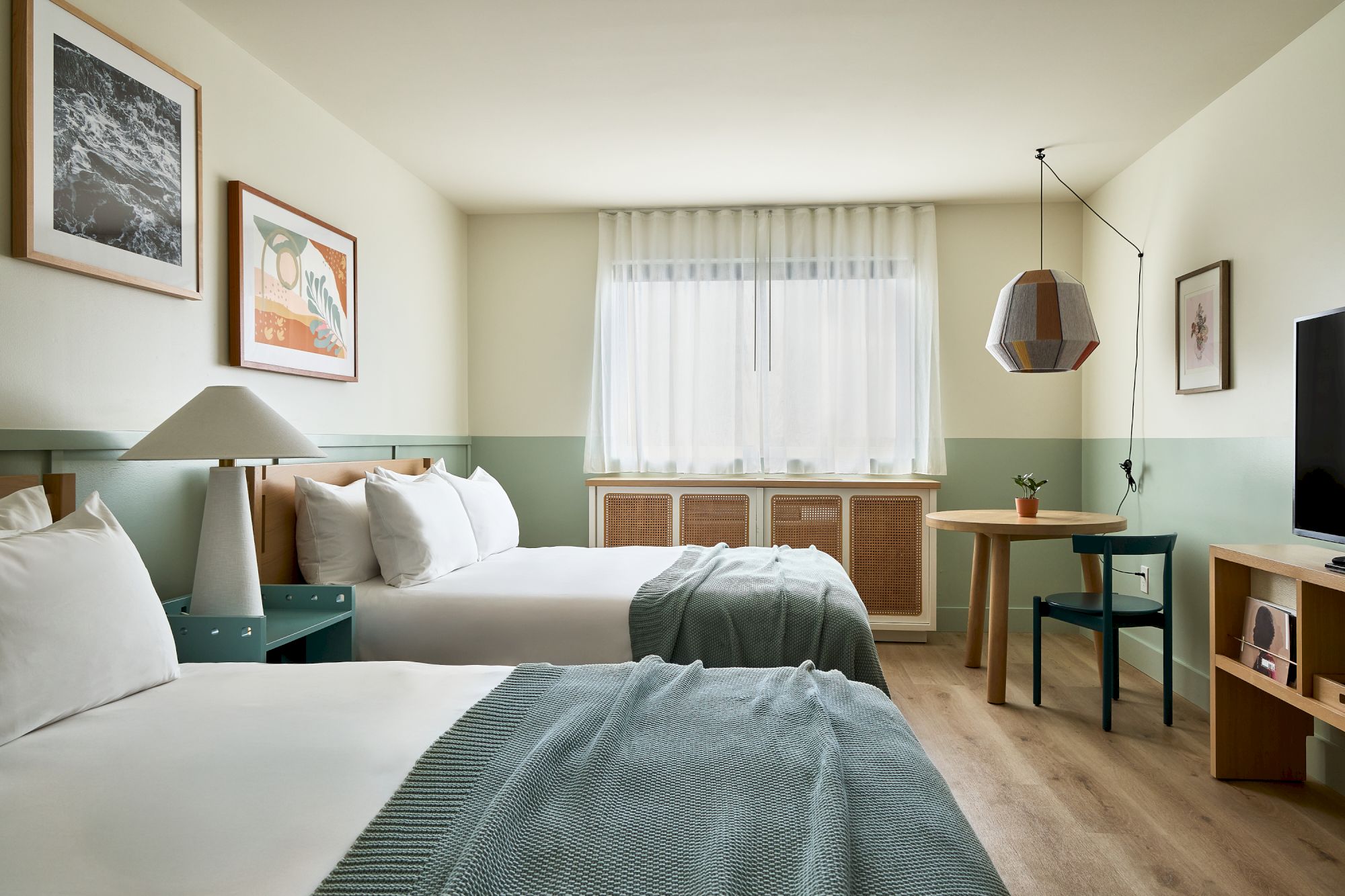 A tidy hotel room with two beds, teal accents, framed art, a window with sheer curtains, a desk, and a flat-screen TV.