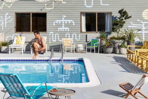 A person sits near a pool surrounded by colorful chairs and modern decor, with abstract designs on the building wall in the background.