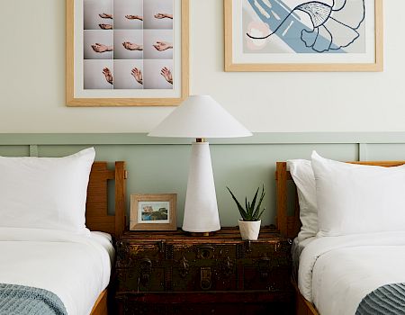 Bright bedroom scene with two framed prints, a white table lamp, a potted aloe plant, and pillows beside a trunk.