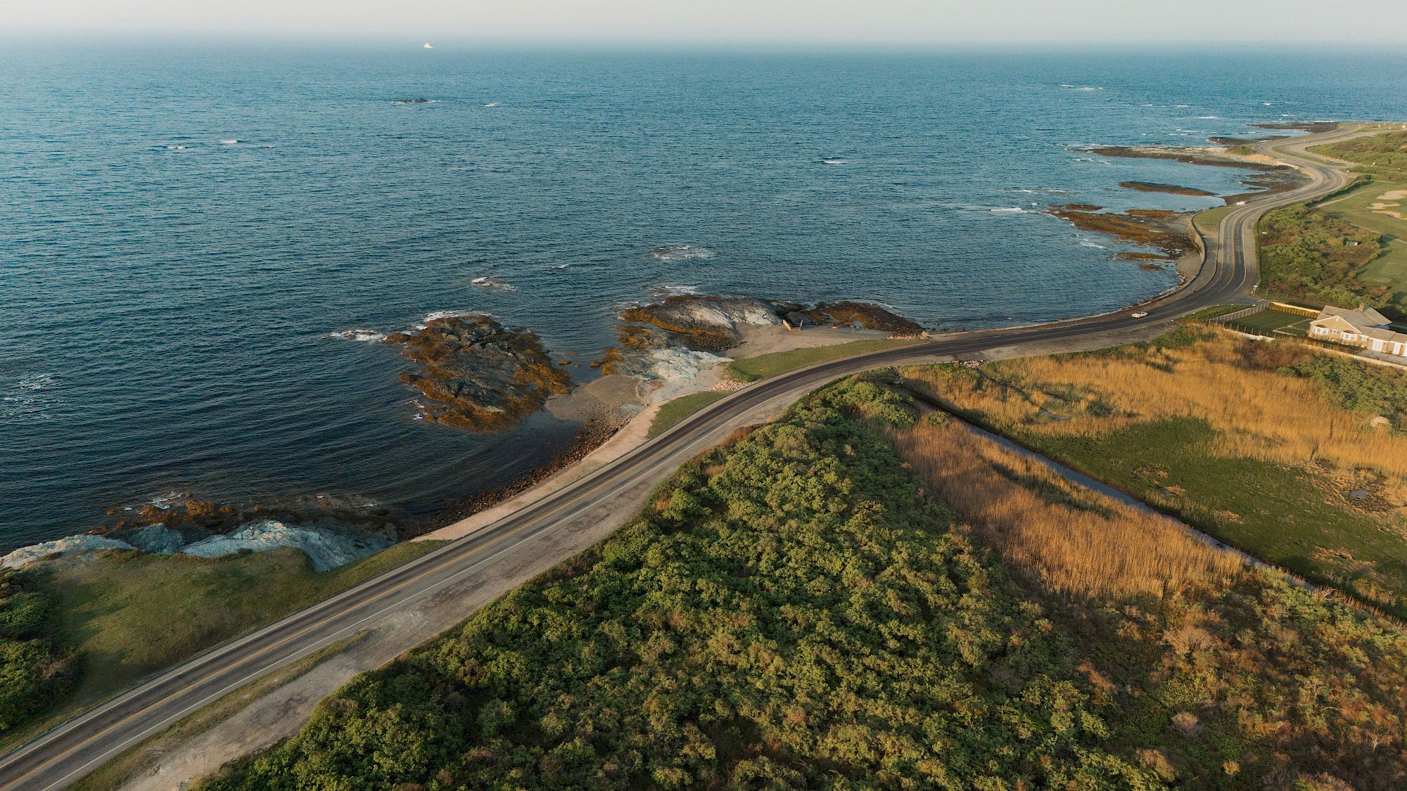 An aerial view of a winding coastal road along rocky shoreline and beaches beside the blue ocean, with grassy cliffs in view.