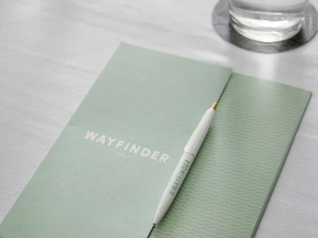 The image shows a green "WAYFINDER" notebook, a white pen with the same branding, and a glass of water on a white surface.