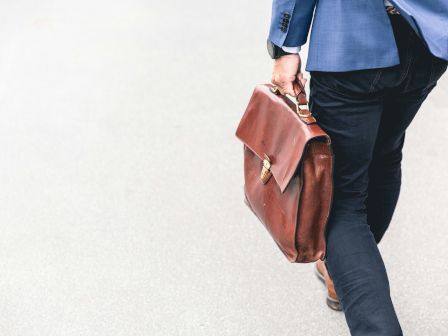 A person in business attire carries a brown leather briefcase while walking on a light-colored surface.
