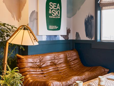 Cozy lounge with a brown leather sofa and plants, featuring a hanging SEA & SKI suntan lotion bottle poster against a wall mural.