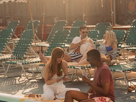 A sunny pool party with people lounging on chairs under striped umbrellas and string lights, chatting in the warm evening.